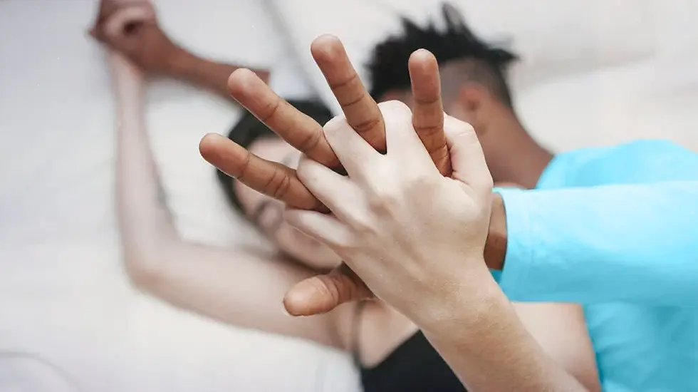 Hands interlock over a blurred background of a couple lying close together. The mood is relaxed and intimate, with soft lighting.
