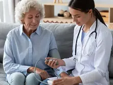 Elderly woman and doctor on a couch, checking blood pressure. Doctor in white coat, stethoscope; calm, professional setting.