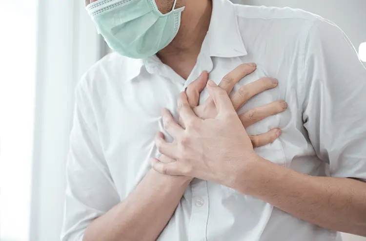 Person in a white shirt and face mask clutching chest with both hands, suggesting discomfort or pain, standing against a bright background.