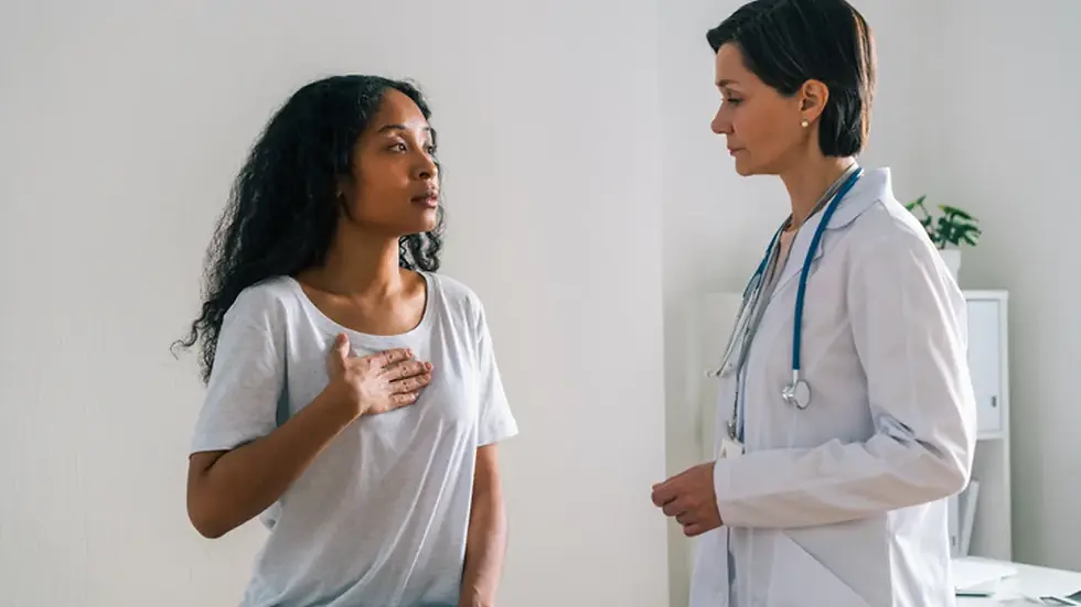 A woman in a gray shirt gestures toward her chest, speaking to a doctor in a white coat with a stethoscope. Neutral-toned room.