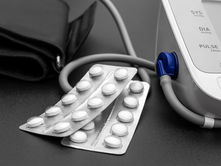 Blood pressure monitor and pill blisters on a black surface. Display shows SYS, DIA, PULSE labels. Blue and white device, clinical setting.