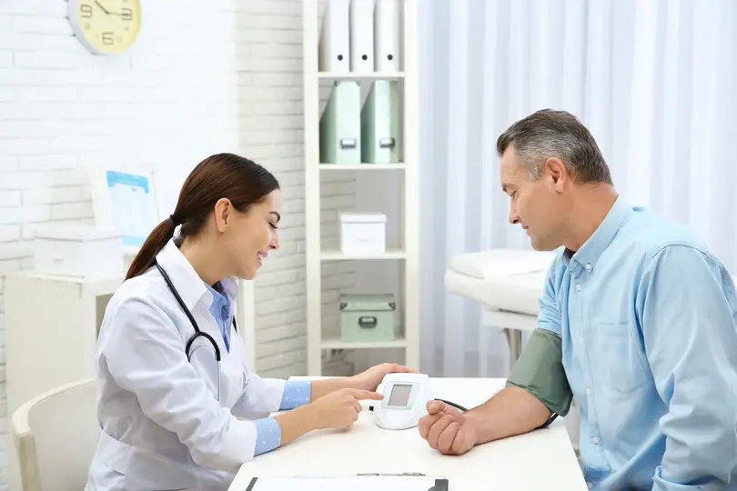 Doctor smiles while checking a man's blood pressure in a bright clinic. White walls, medical equipment, and a clock visible.