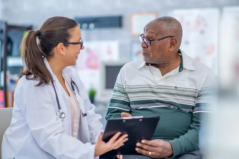 Doctor and elderly man talking in a clinic. The doctor holds a clipboard, both seem engaged. Medical posters visible in the background.