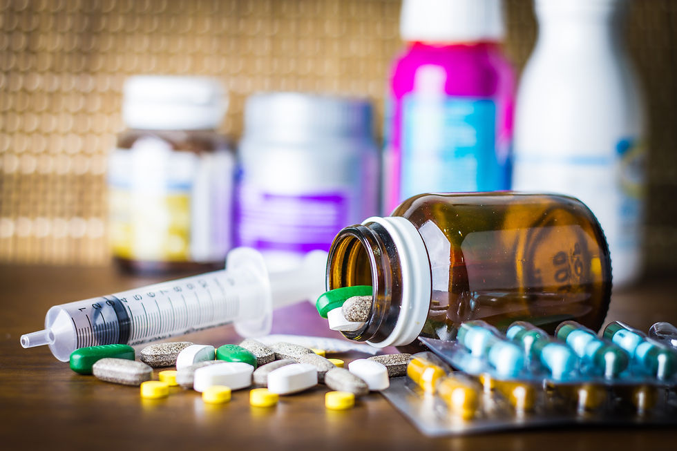 Pills spill from a brown bottle onto a table with a syringe nearby. Blurred colorful medicine bottles in the background.