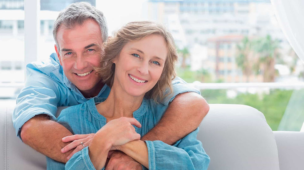 Smiling couple in blue shirts cuddling on a sofa with a bright window view behind them, conveying warmth and happiness.