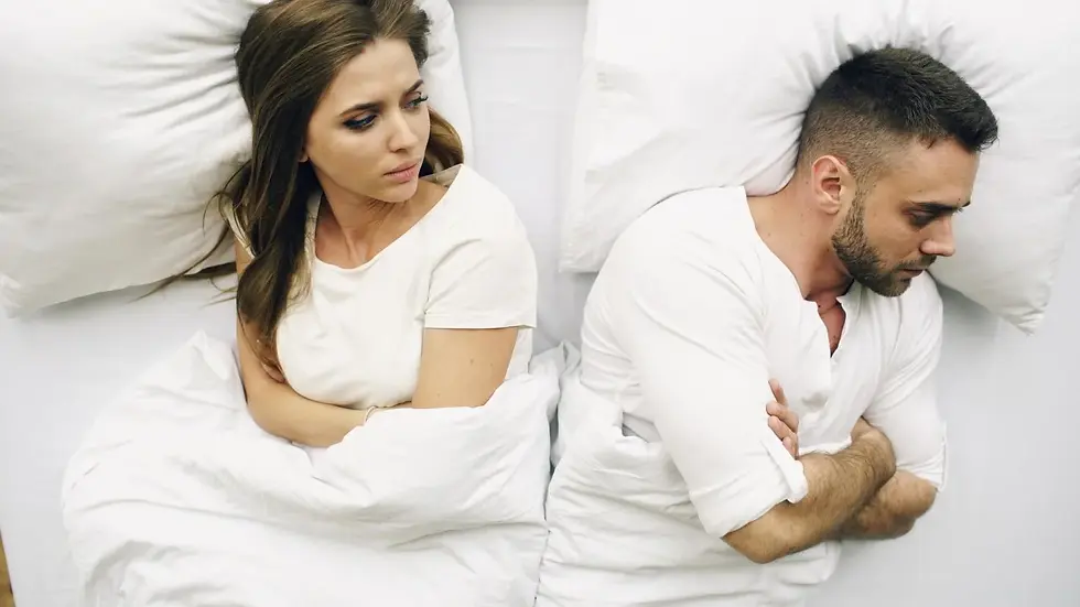 A man and woman lie back-to-back in bed, arms crossed, wearing white shirts. The mood is tense. White pillows and bedding surround them.