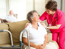 A nurse in pink scrubs comforts an elderly woman sitting on a sofa. A man reads in the background. Indoor setting with soft lighting.