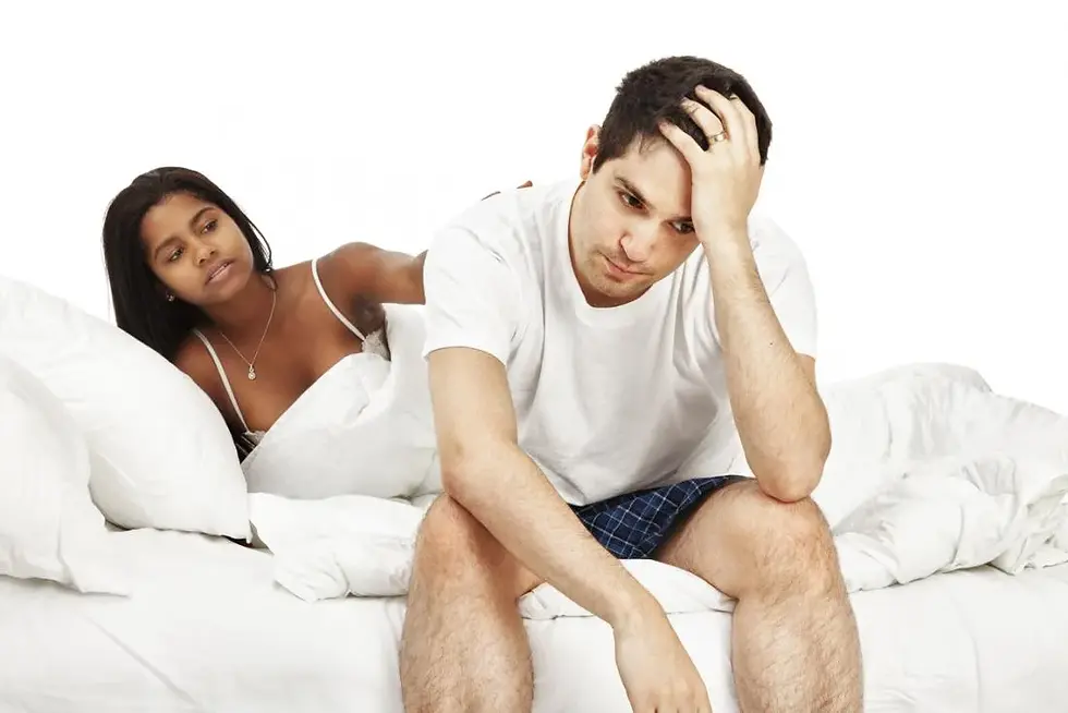Man in white t-shirt looks distressed, sitting on bed. Woman in white tank top reclines behind him, appearing concerned. White bedding.