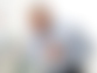 Elderly man clutching chest in pain in a bright room with soft furnishings. He appears distressed, wearing a light blue shirt.