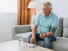 Elderly man in a turquoise shirt checks blood pressure on a sofa. Table with pills, water, and device. Bright, cozy living room. Calm mood.