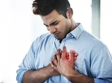 Man in a blue shirt clutches his chest in pain, eyes closed, against a bright, blurred background, conveying discomfort and concern.