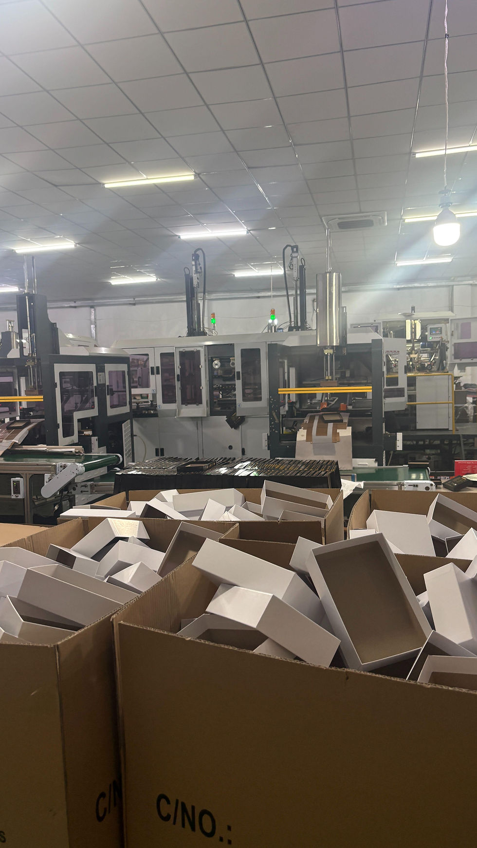 Open rectangular box components in cartons beside equipment in a packaging workshop.