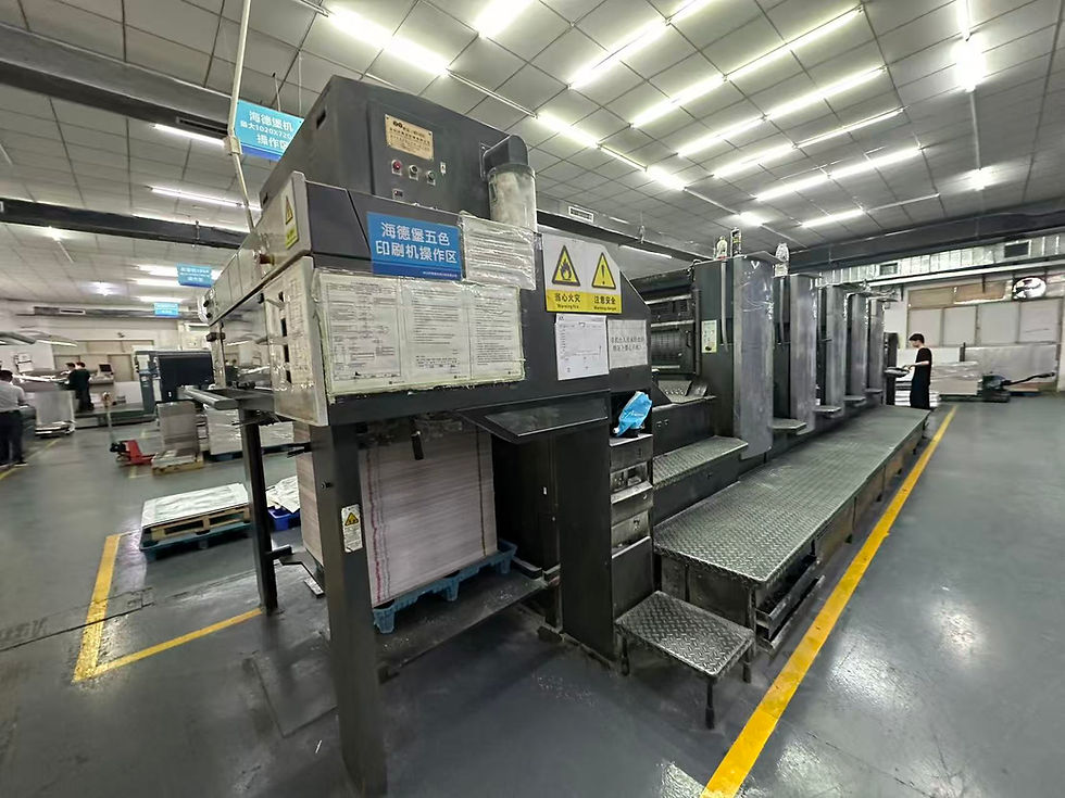 Large industrial printing press in a factory, with Chinese safety signs, paper stacks, and workers nearby under bright lights.