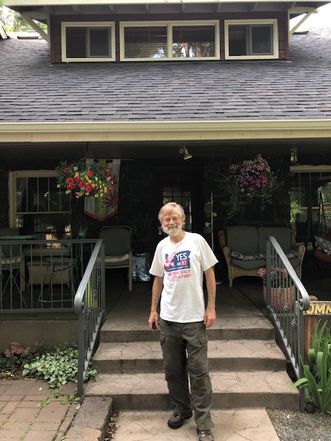 A bearded, smiling man stands in front of a country-style porch.