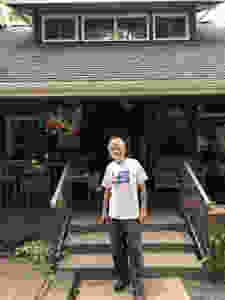 A bearded, smiling man stands in front of a country-style porch.