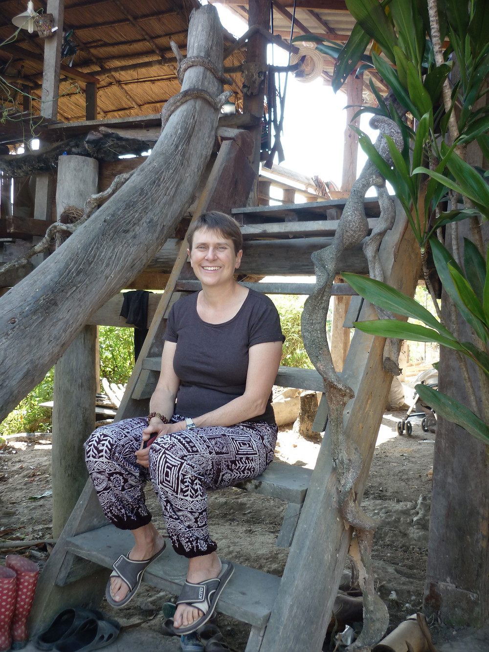 I am sitting on steep, rustic front steps. A tree trunk serves as a handrail. Over the house is a thatch roof, and we can see out the back of the house because there is no wall.