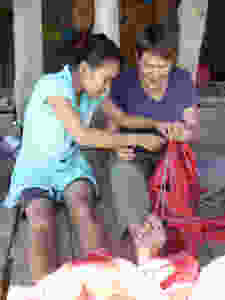 A Thai woman sits beside Jeannine on the floor, showing her how to sew a seam on a traditional piece of clothing. A string looped around the toe helps to keep tension in the fabri.