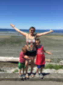 A woman in her thirties holding her arms out joyfully as her two boys hang on to her. There is a beach in the background.