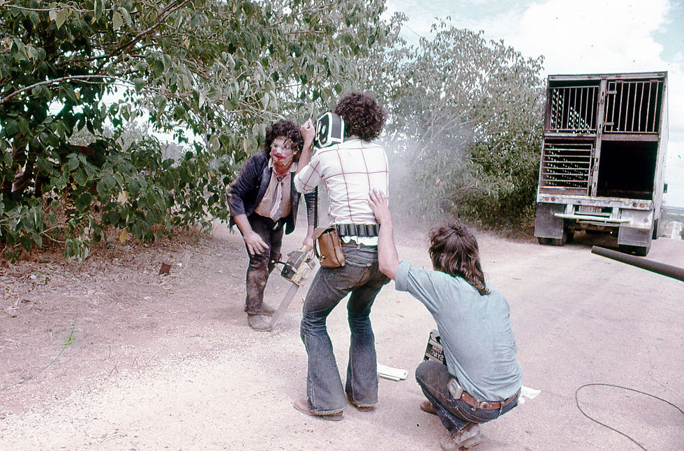 Three people in a dusty outdoor setting. One uses a chainsaw near a film camera, with trees and a truck in the background. Casual attire.