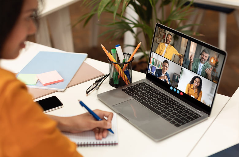 A woman is sitting at a desk, taking notes in a notebook while participating in a video co
