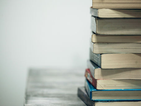 A stack of books on a wooden table against a plain light gray background. The books have various colored covers and visible wear.