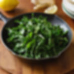 Fresh greens in a black skillet on a wooden surface, with a lemon, garlic, and plates in the background. Bright, natural setting.