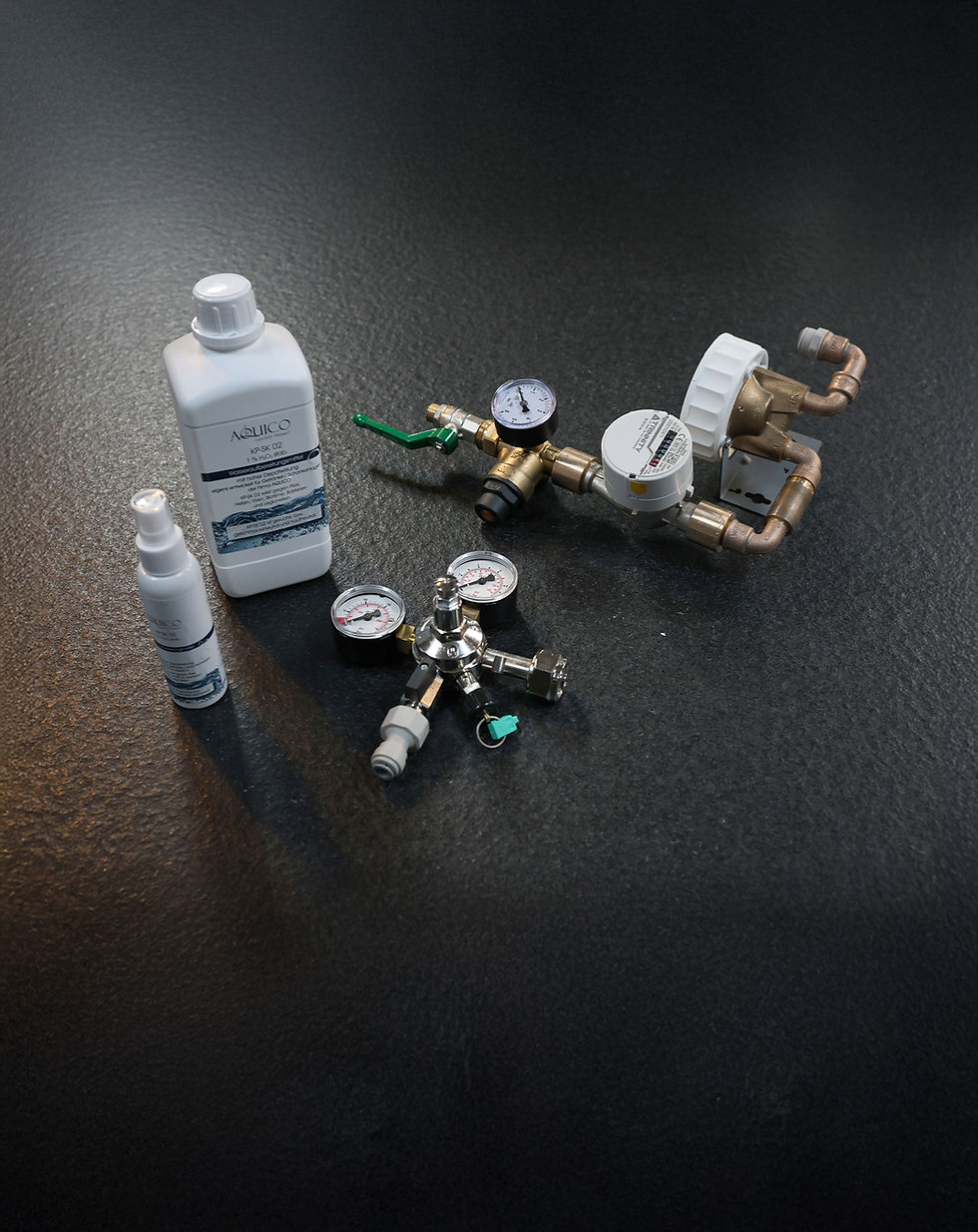 Zubehör-Set für Wasserspender von Aquico, auf dunkler strukturierter Oberfläche. Enthalten sind ein weißer Kanister und eine kleine Flasche mit Aquico-Logo, ein Druckminderer mit zwei Manometern, ein CO₂-Druckregler mit drei Anschlüssen und weitere Mess- und Ventilelemente aus Metall und Kunststoff.
