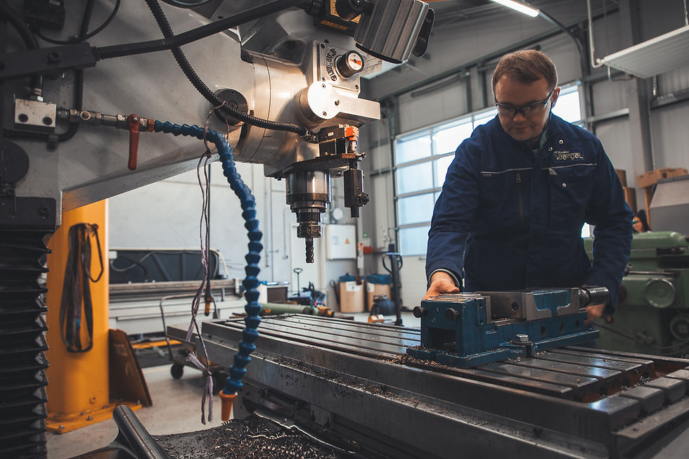 Ein Mitarbeiter in blauer Arbeitskleidung mit dem Firmenlogo „Pengel“ steht an einer großen Fräsmaschine. Er trägt Schutzbrille und richtet mit der Hand ein Werkstück oder einen Schraubstock auf dem Arbeitstisch der Maschine aus. Die Fräse ist mit einem Spiralbohrer bestückt, Kühlmittelschläuche hängen seitlich herab. Metallspäne liegen auf dem Maschinentisch, was auf laufende oder kürzlich durchgeführte Bearbeitung hindeutet. Die Werkstatt ist hell, modern und gut ausgestattet, im Hintergrund sind Fenster, Maschinen und Werkbänke sichtbar. Das Bild vermittelt ein professionelles Umfeld in der mechanischen Fertigung oder Metallverarbeitung.