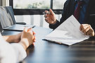 Zwei Personen sitzen sich in einem modernen Büro an einem Tisch gegenüber. Eine Person, gekleidet im Anzug mit Krawatte, hält ein Blatt Papier mit Geschäftsdaten und gestikuliert mit einem Kugelschreiber. Die andere Person hört aufmerksam zu und hat die Hände gefaltet. Im Hintergrund steht ein aufgeklappter Laptop auf dem Tisch.