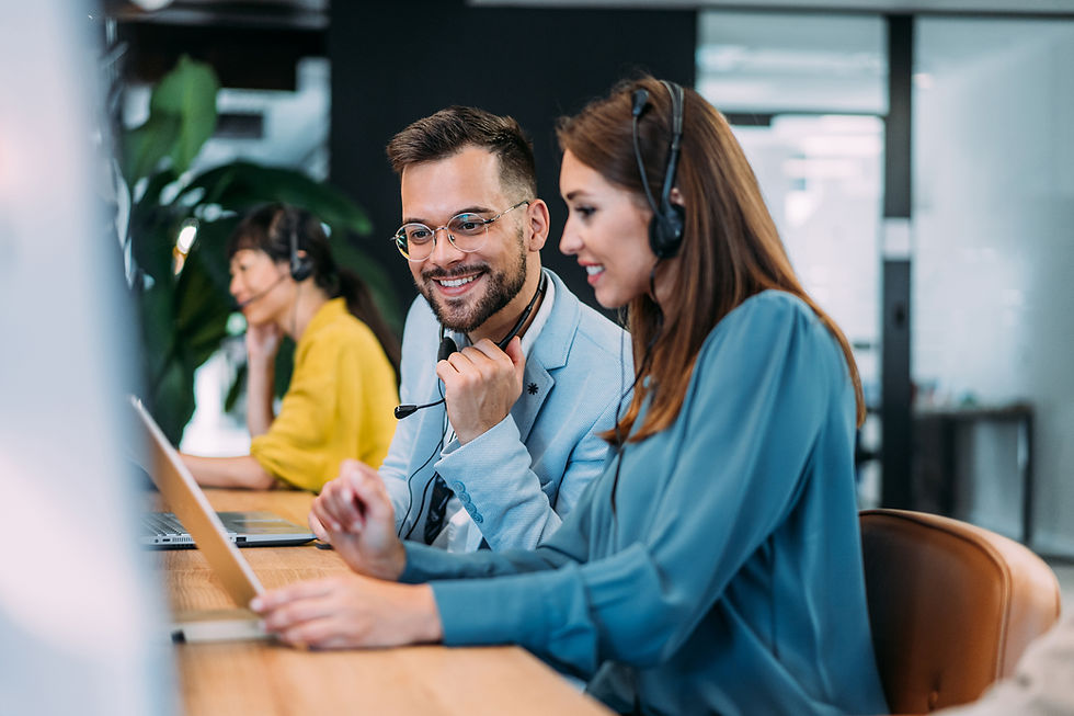 Zwei freundliche Kundenservice-Mitarbeiter in einem modernen Büro, beide mit Headset und vor Laptops, arbeiten gemeinsam und lächeln in die Kamera, während im Hintergrund eine Kollegin telefoniert.