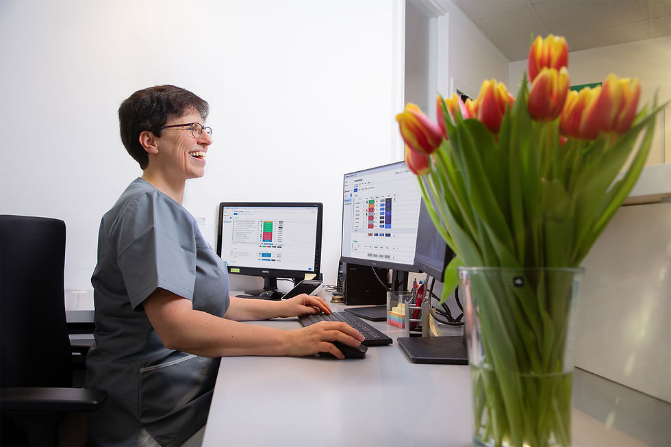 Mitarbeiterin einer Zahnarztpraxis sitzt lachend an der Rezeption vor zwei Monitoren mit geöffneter Praxissoftware. Im Vordergrund steht ein Strauß rot-gelber Tulpen in einer Glasvase.