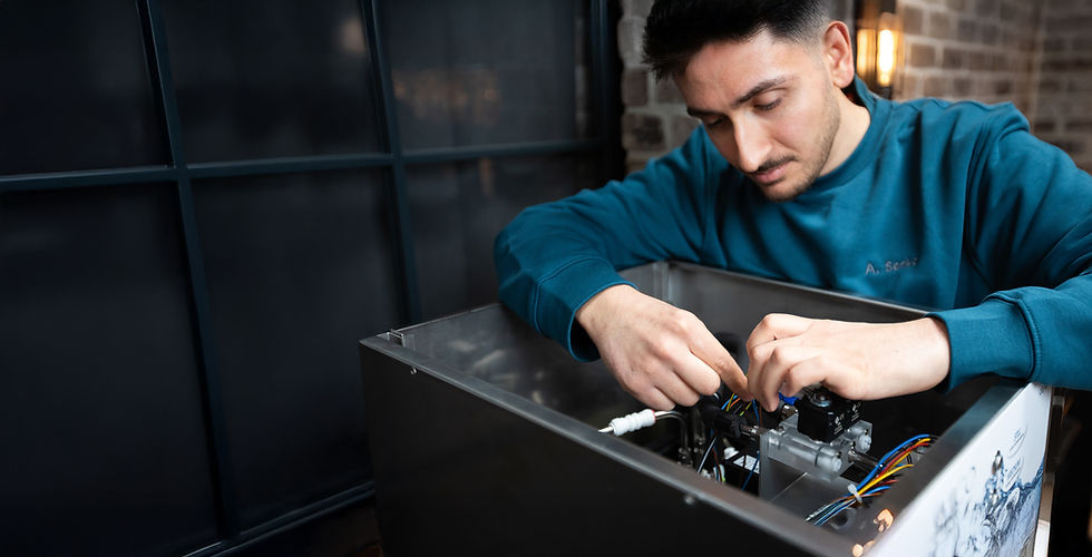 Ein Techniker in einem türkisfarbenen Sweatshirt arbeitet konzentriert am Inneren eines Wasserspenders. Er überprüft Kabel und Bauteile in einem modernen Raum mit Glaswand und Backsteinoptik.