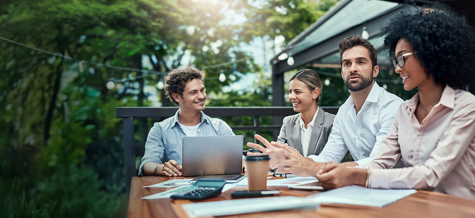 Vier Personen, zwei Männer und zwei Frauen, sitzen an einem Holztisch im Freien und führen ein Gespräch. Auf dem Tisch liegen Laptops, Dokumente, ein Taschenrechner und Kaffeebecher. Die Gruppe wirkt konzentriert, während sie über geschäftliche Themen spricht, mit grünem Hintergrund aus Bäumen und sanftem Sonnenlicht.