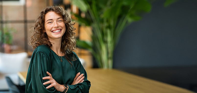 Junge Frau mit lockigen Haaren und grünem Oberteil steht lächelnd mit verschränkten Armen in einem modernen Büro oder Arbeitsraum. Im Hintergrund sind unscharf Pflanzen, ein großer Tisch und eine helle Sitzecke zu sehen, was dem Bild eine freundliche und professionelle Atmosphäre verleiht.