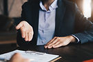 Nahaufnahme einer Person im Anzug während eines Gesprächs am Tisch. Eine geöffnete Hand ist gestisch erhoben, die andere ruht auf dem Tisch. Vor der Person liegt ein Vertrag.