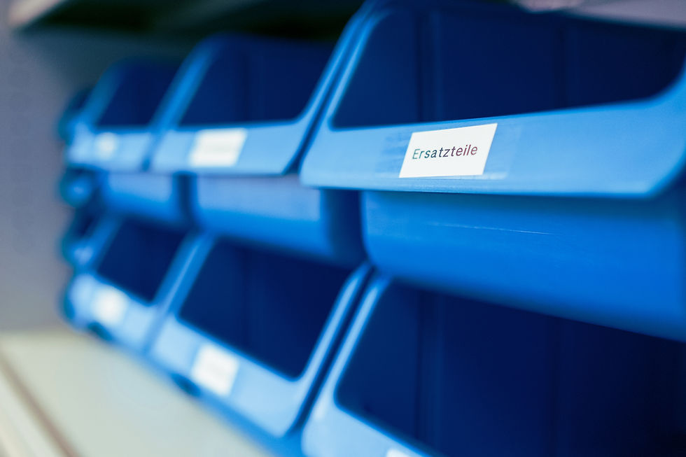 Mehrere leere, blaue Lagerboxen in einem Regal, eine davon mit einem weißen Etikett „Ersatzteile“ beschriftet.