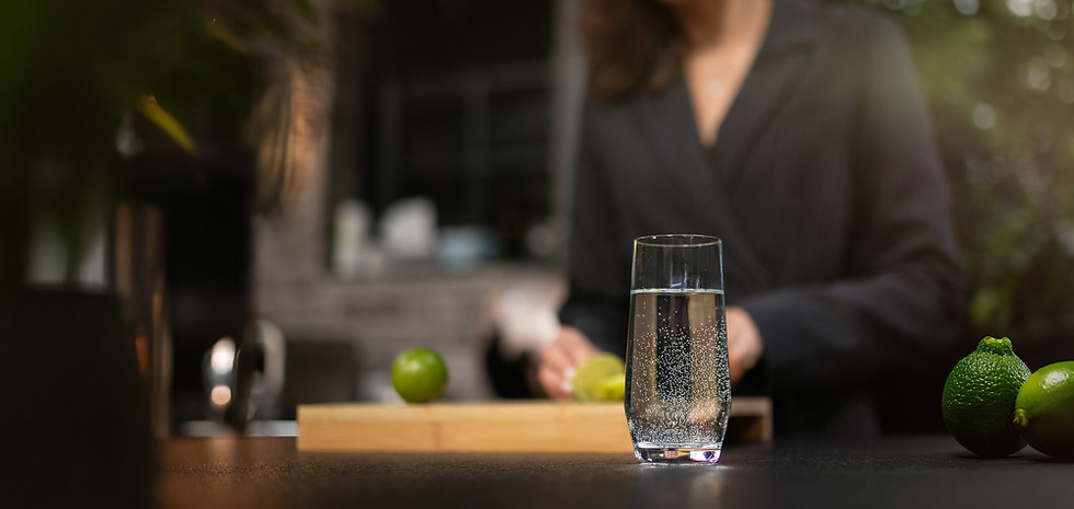 Ein Glas mit sprudelndem Wasser steht im Vordergrund auf einer dunklen Tischplatte. Daneben liegen zwei frische, grüne Limetten. Im Hintergrund unscharf eine Frau in Business-Kleidung, die an einem Schneidebrett weitere Limetten schneidet. Die Szenerie ist warm beleuchtet, mit Pflanzen und weicher Tiefenschärfe, was eine ruhige und frische Atmosphäre vermittelt.