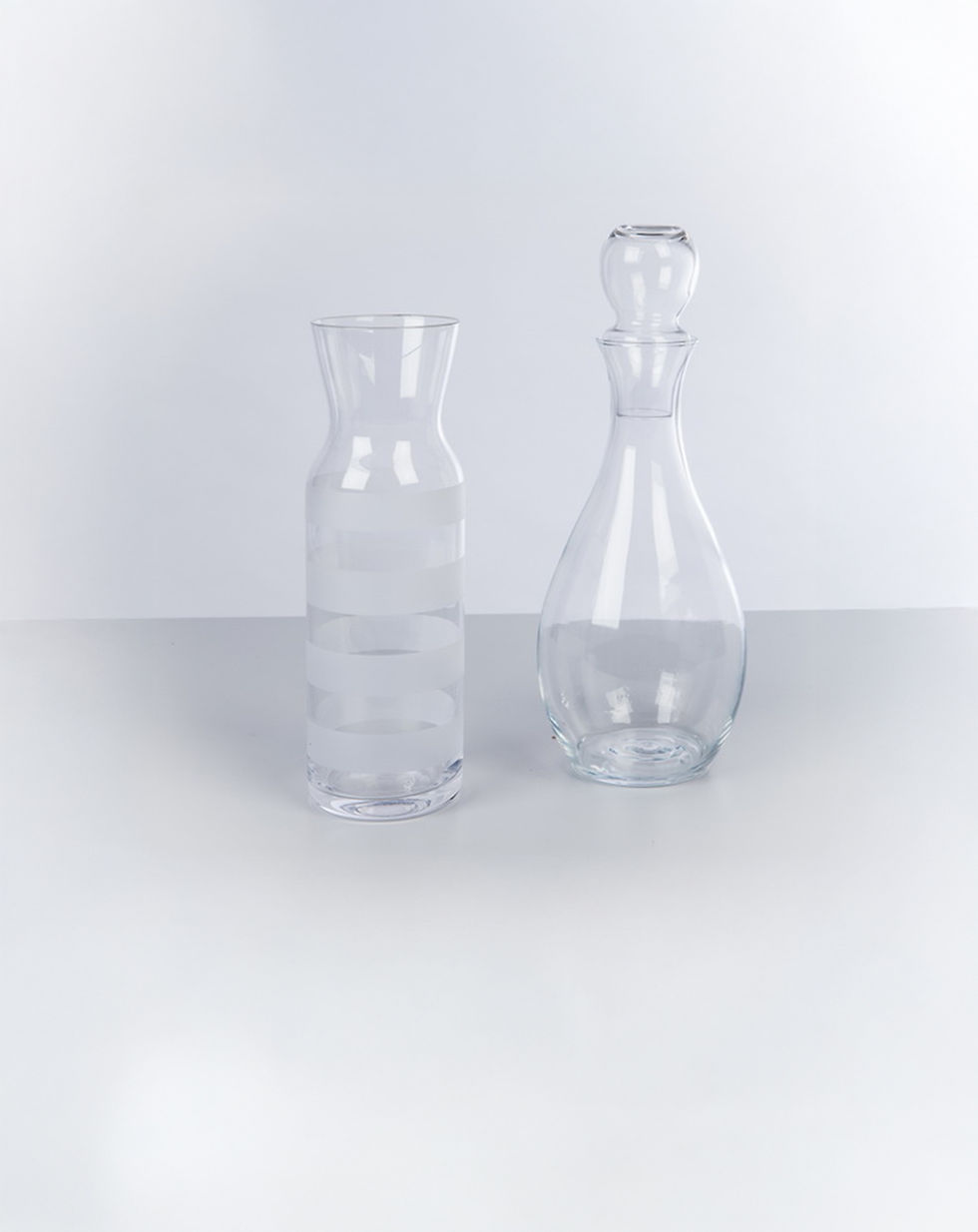 Zwei transparente Glas-Karaffen auf weißem Hintergrund. Links eine zylindrische Karaffe mit leicht ausgestelltem Hals und matten Zierstreifen, rechts eine bauchige Karaffe mit Stöpsel und elegantem, tropfenförmigem Design.