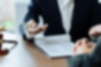 Zwei Personen sitzen an einem Tisch und führen ein Beratungsgespräch. Im Vordergrund liegt ein aufgeschlagener Vertrag oder ein juristisches Dokument. Eine der Personen trägt einen dunklen Anzug und hält einen Kugelschreiber in der Hand, als wolle sie etwas erklären oder zeigen. Die andere Person hat die Hände gefaltet. Links am Bildrand ist eine goldene Justitia-Waage sichtbar – Symbol für Recht und Gerechtigkeit.