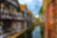 Canal between half-timbered houses and a brick wall, with flower boxes, moored boats, and a cloudy sky.