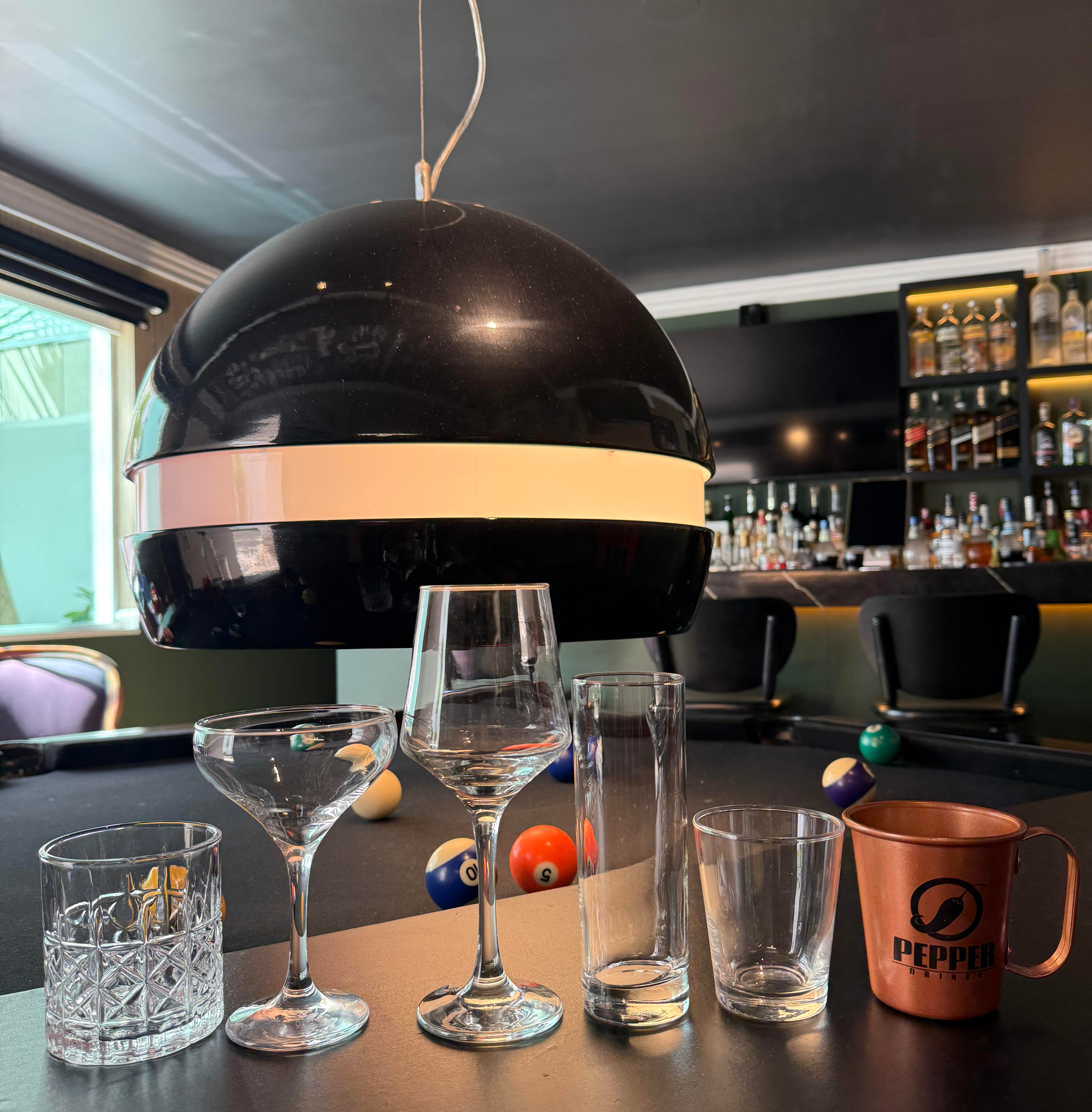 Mesa de bar com copos vazios e caneca cobre Pepper Drinks; ao fundo, mesa de sinuca, garrafas e iluminação escura.
