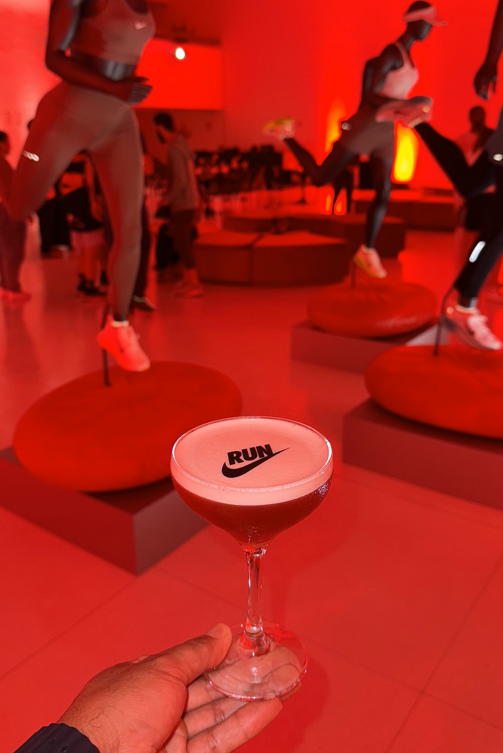 Mão segura taça de coquetel com RUN, em sala vermelha com manequins de corredores e clima de evento esportivo.