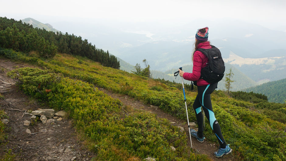 Woman hiking uphill on a mountain trail using trekking poles, wearing a backpack in cold, foggy weather with scenic valley views