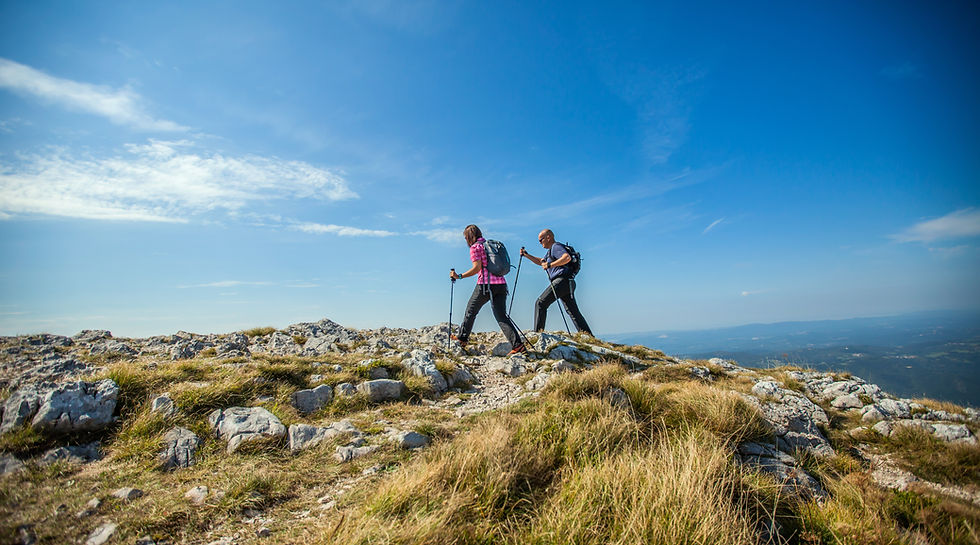 Two hikers using trekking poles walking across a rocky mountain plateau under a clear blue sky with scenic landscape views