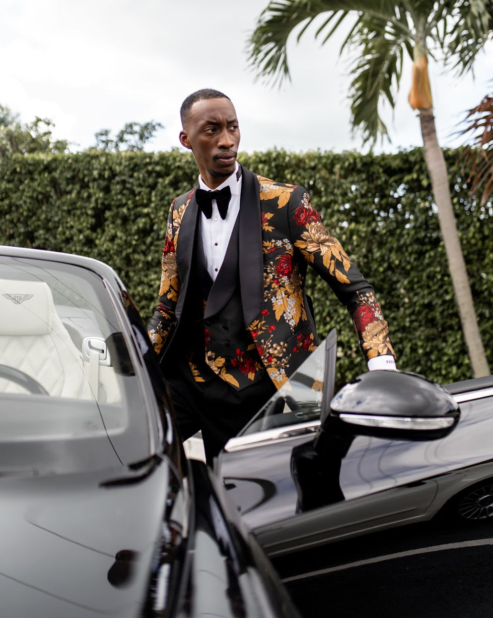 a man in a tuxedo stands next to a bentley car