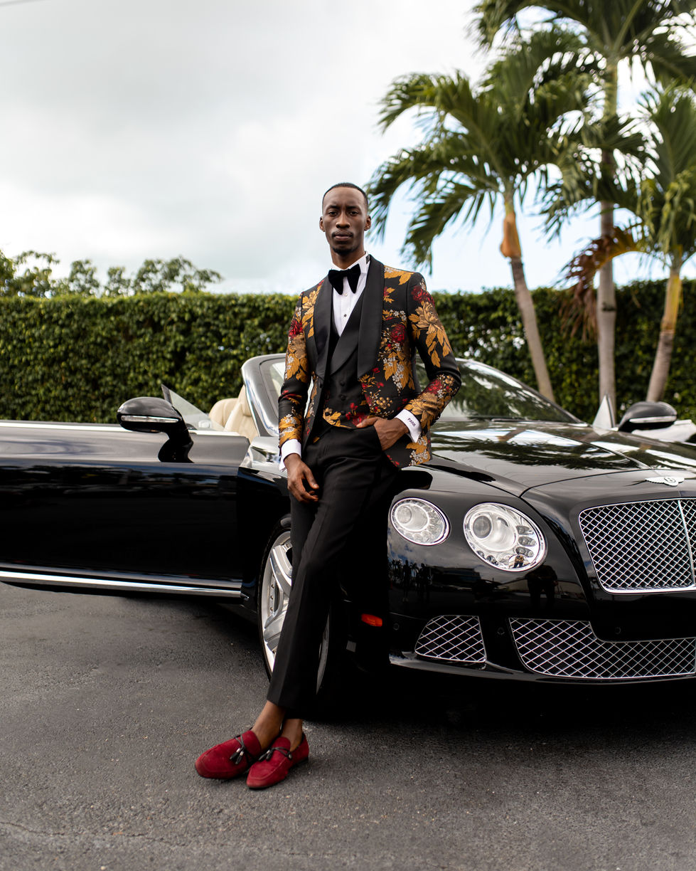 a man in a tuxedo leans against a black bentley