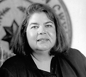 Wilma Mankiller, smiling, in black and white. Behind her is a carpet hung on the wall.