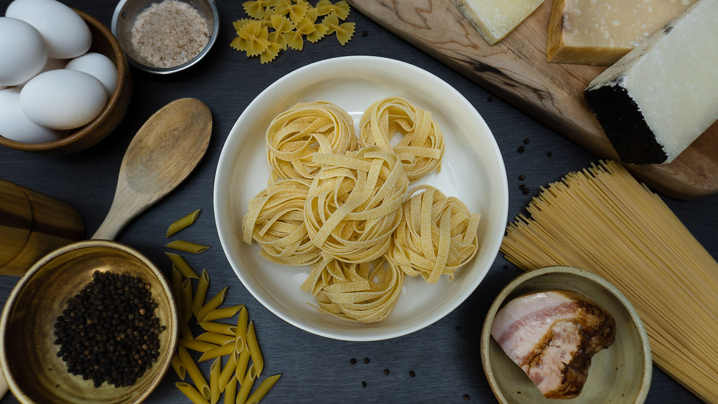 Pastabasta hero - ingredients on dark surface carbonara components laid out on table dry pasta guanciale eggs pepper salt cheese in bowls