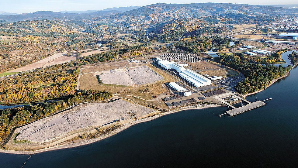 Aerial view of the proposed Kalama methanol facility site at the Port of Kalama, showing large white industrial buildings and empty lots beside the Columbia River, surrounded by wooded hills with autumn foliage.