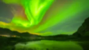 Grün-rote Polarlichter tanzen über einer Bucht der Lofoten, reflektiert im ruhigen Wasser, umgeben von schroffen Bergen.
